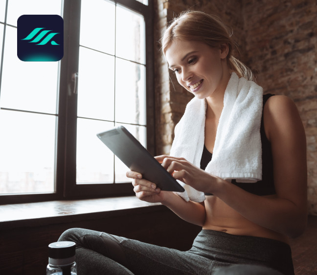 Uśmiechnięta kobieta w sportowym stroju i ręcznikiem na szyi korzysta z tabletu.