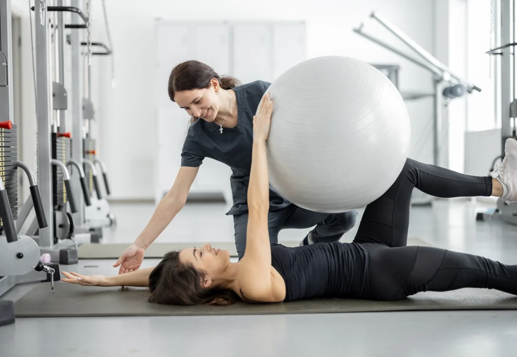 Fizjoterapeutka w szarym uniformie pomaga uśmiechniętej pacjentce w ćwiczeniach z dużą piłką rehabilitacyjną w jasnej sali do ćwiczeń. Pacjentka leży na plecach na macie, trzymając piłkę nogami, a fizjoterapeutka pochyla się nad nią, udzielając wskazówek.