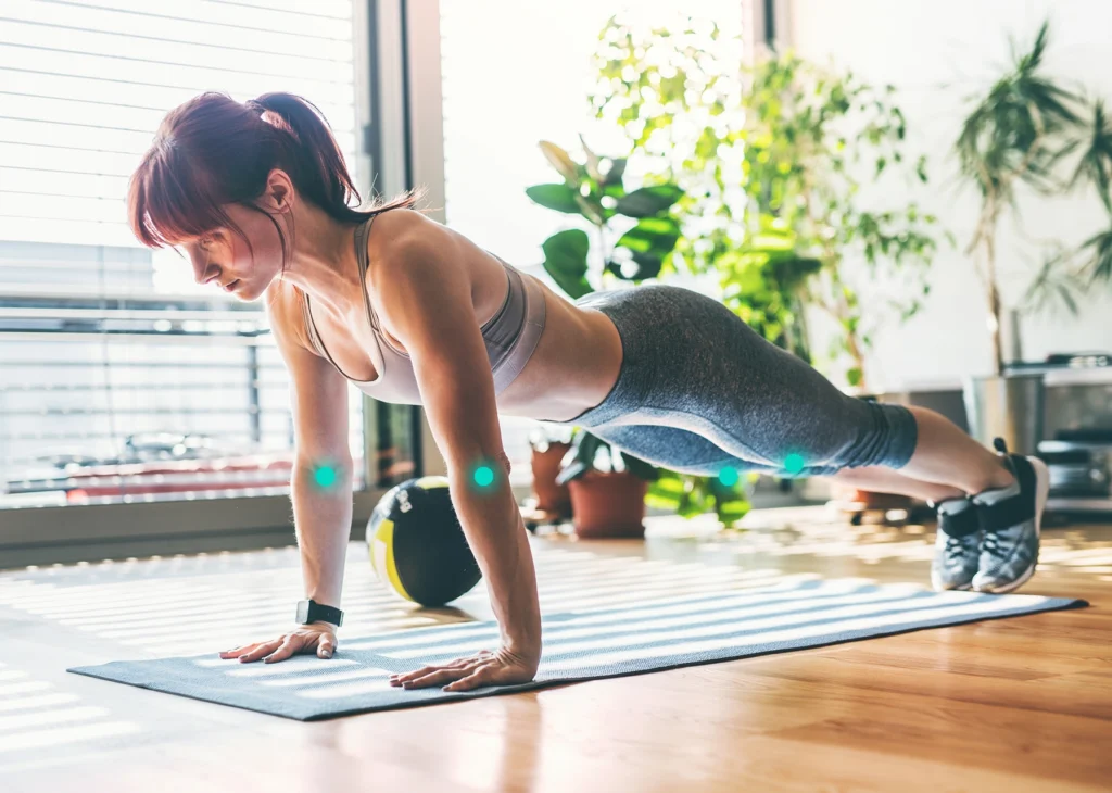 Kobieta w sportowym stroju wykonuje plank na macie w jasnym pomieszczeniu z roślinami i sprzętem do ćwiczeń w tle. Na jej łokciach i kolanach zaznaczone są punkty symbolizujące czujniki ruchu.